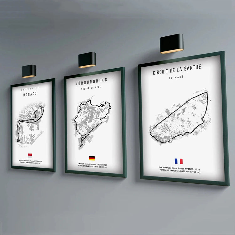 Three framed minimalist F1 formula one track map prints displayed on a grey wall, each illuminated by a small picture light, featuring black line illustrations of geographic regions with country labels and flags beneath.